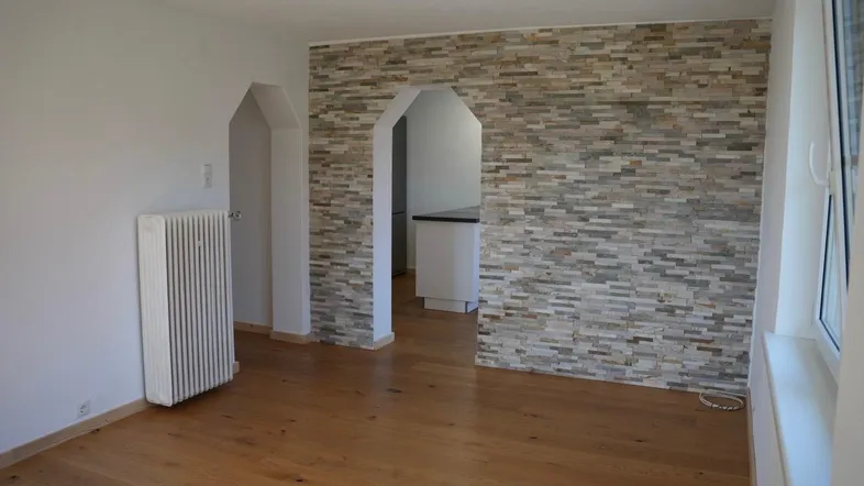 Heller Wohnbereich mit Holzboden und dekorativer Steinwand, offen zu einem weiteren Raum.