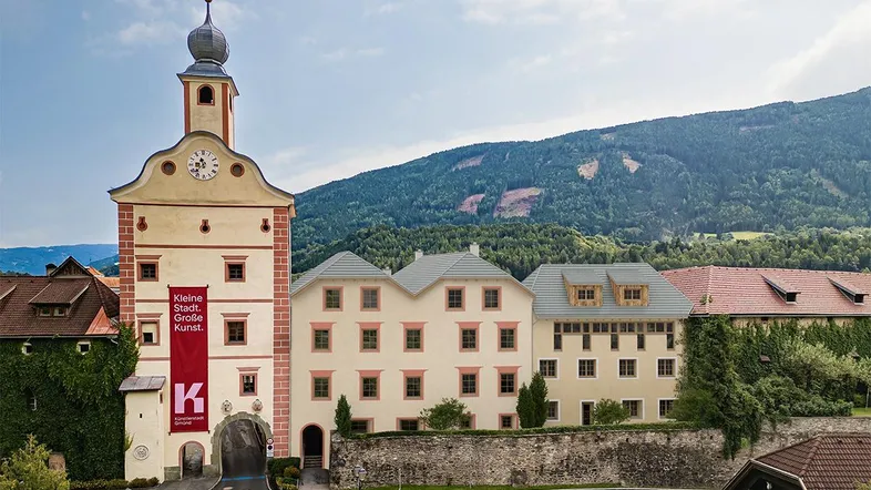Historisches Gebäude mit Uhrturm und traditioneller Architektur, umgeben von grünen Bergen unter blauem Himmel.