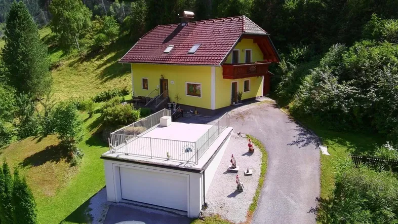 Luftaufnahme eines gelben Hauses mit rotem Dach, Garage und Dachterrasse in grüner Berglandschaft.