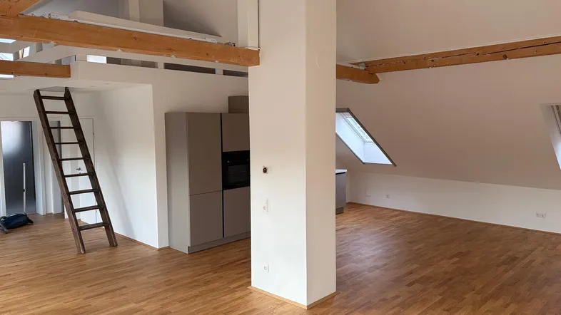 Heller offener Wohnbereich mit Holzbalken, Dachschrägen und integrierter Küchenzeile, ausgestattet mit Parkettboden.