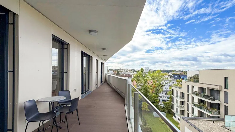 Langer Balkon mit Holzboden, Glasgeländer und weitem Stadtblick unter blauem Himmel.