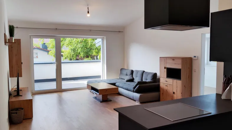 Heller Wohnbereich mit Holzboden, Zugang zum Balkon und Blick auf die offene Küche.