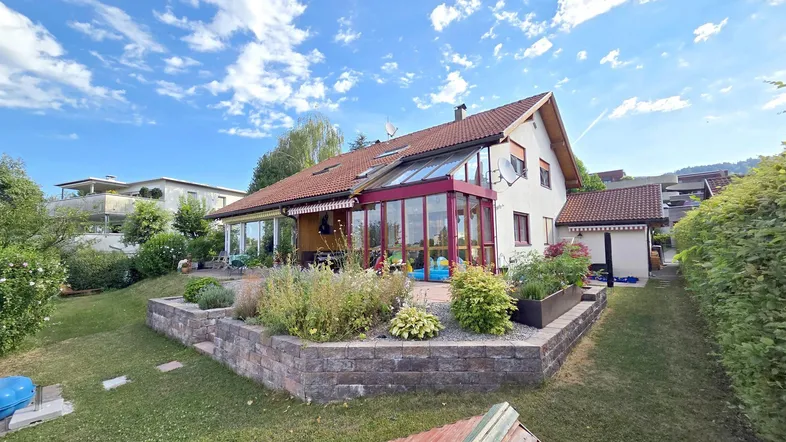 Hausansicht von hinten mit großem Garten, Wintergarten und gepflegten Beeten unter blauem Himmel.