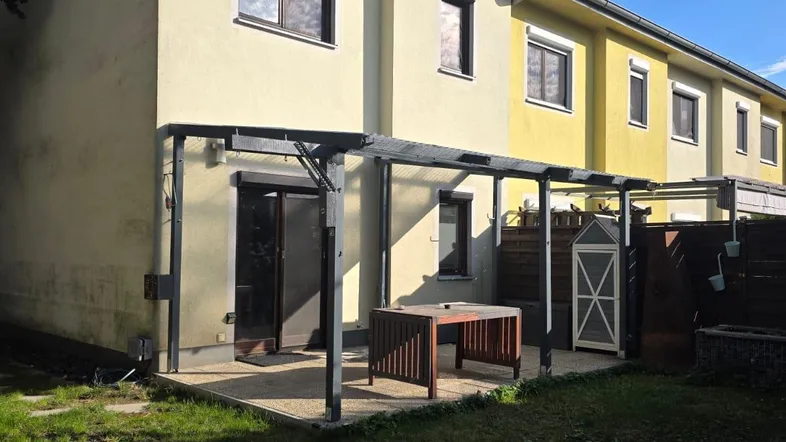 Hinterhofansicht eines Reihenhauses mit überdachter Terrasse und Rasenfläche unter blauem Himmel.