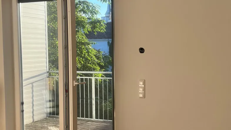 Helles Zimmer mit Holzboden und Zugang zu einem Balkon, der einen Blick ins Grüne bietet.
