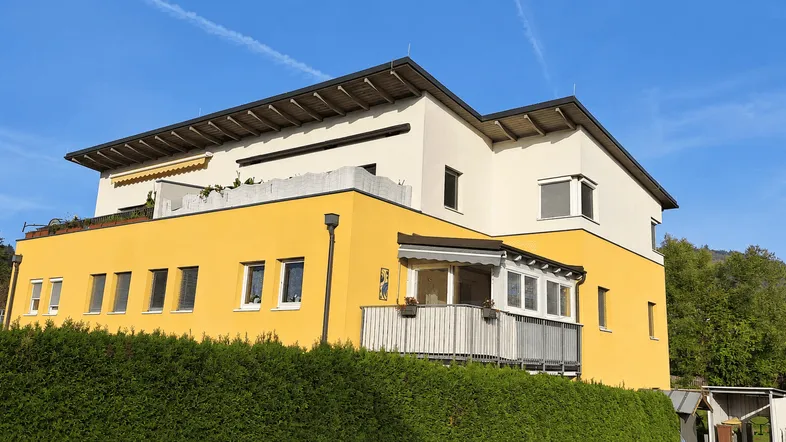 Modernes Mehrfamilienhaus mit gelber Fassade, Balkonen und Dachterrasse unter blauem Himmel.
