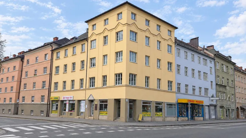 Gelbes Eckgebäude mit mehreren Stockwerken und Geschäften im Erdgeschoss an einer belebten Kreuzung.