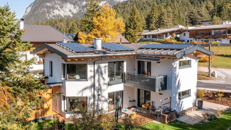 Modernes Haus mit Solaranlage, Terrasse und Balkon, umgeben von Natur und Bergen.