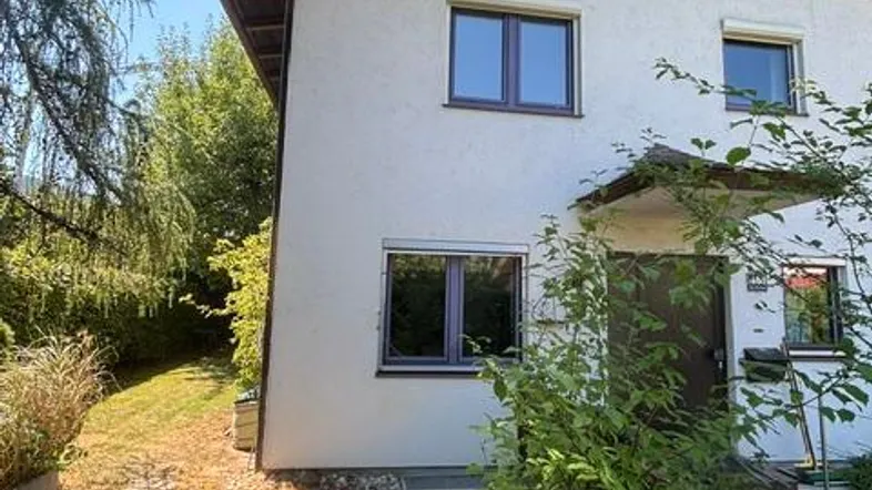 Vorderansicht eines Hauses mit weißer Fassade, braunem Dachüberstand und grüner Umgebung.