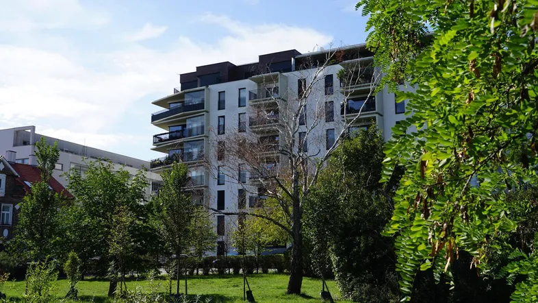 Modernes Mehrfamilienhaus mit Balkonen, umgeben von Bäumen und Grünflächen unter blauem Himmel.