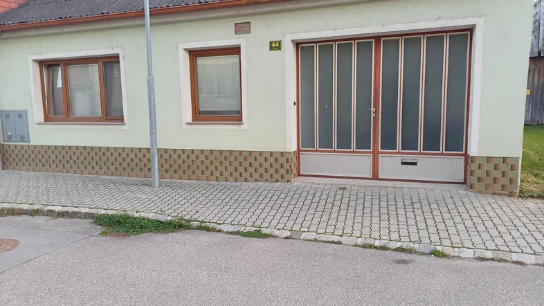Fassade eines Hauses mit Garagentor und kleinen Fenstern, umgeben von einem gepflasterten Gehweg.