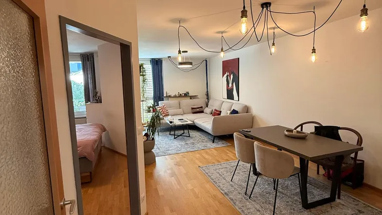 Heller offener Wohn- und Essbereich mit Holzboden, moderner Beleuchtung und Zugang zu einem Schlafzimmer.