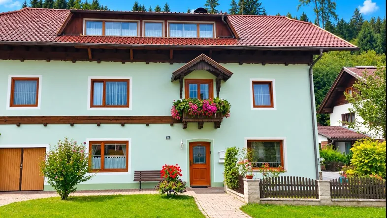 Traditionelles Haus mit hellgrüner Fassade, rotem Ziegeldach und Blumenkästen am Balkon.