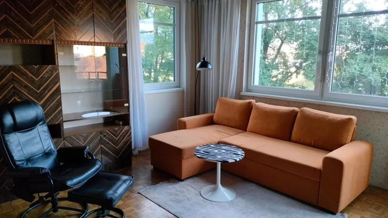 Gemütliches Wohnzimmer mit orangefarbenem Sofa, Holzboden und großen Fenstern mit Blick ins Grüne.