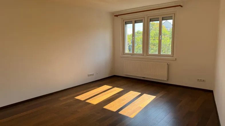 Helles Zimmer mit dunklem Parkettboden und Fenster, das viel Tageslicht hereinlässt.