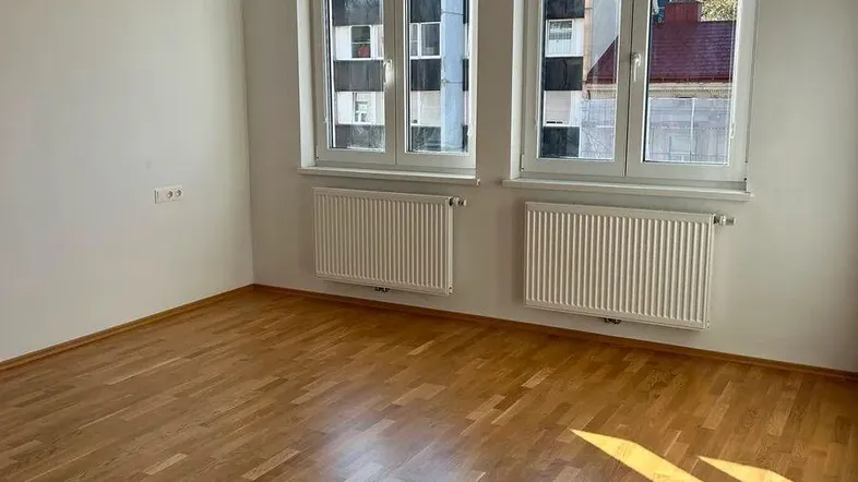 Helles Zimmer mit Parkettboden und zwei Fenstern, die viel Tageslicht hereinlassen.