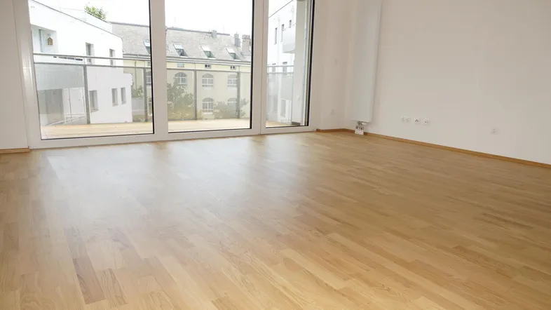 Helles, leeres Zimmer mit Holzboden und großen Fenstern, die auf einen Balkon führen.