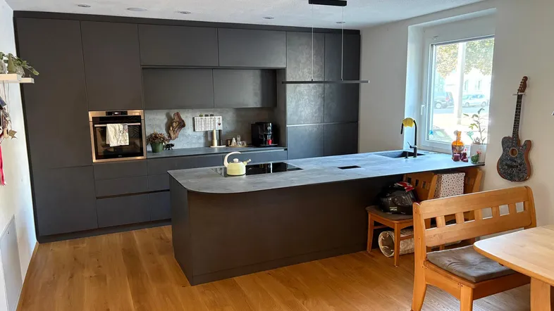 Moderne Küche mit dunklen Fronten, Kochinsel und Holzboden, hell erleuchtet durch ein großes Fenster.