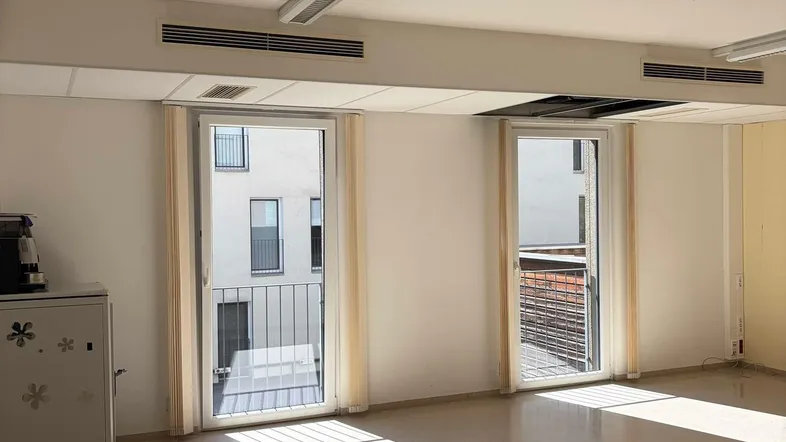 Heller, leerer Raum mit großen Fenstertüren, die viel Tageslicht hereinlassen und einen Blick auf andere Gebäude bieten.
