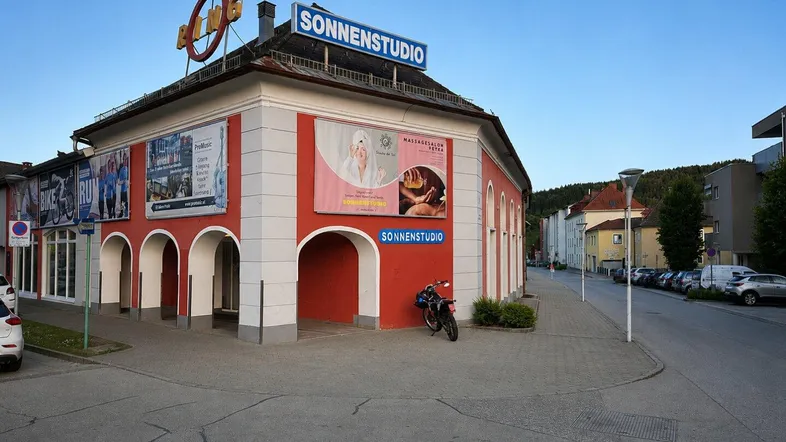 Außenansicht eines Gebäudes mit roten Arkaden und einem "Sonnenstudio"-Schild unter blauem Himmel.