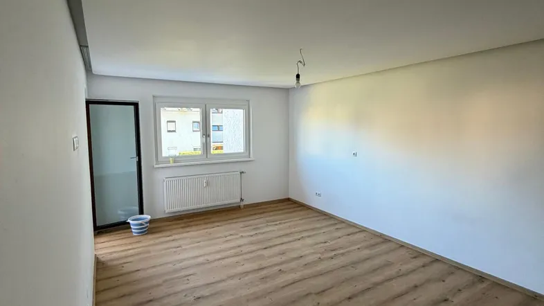 Helles, leeres Zimmer mit neuem Laminatboden und einem Fenster, das Tageslicht hereinlässt.