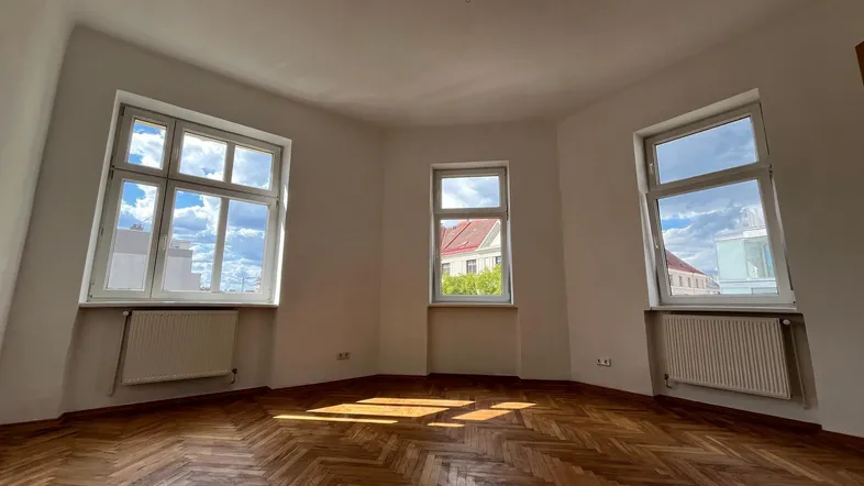 Helles Zimmer mit Fischgrätparkettboden und mehreren Fenstern, die viel Tageslicht hereinlassen.