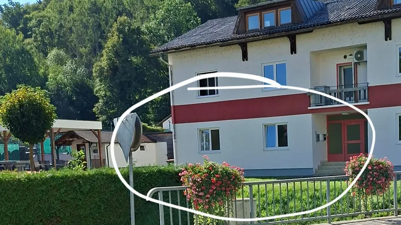 Mehrfamilienhaus mit roter Fassadenakzentuierung, umgeben von viel Grün und einem bewaldeten Hügel.