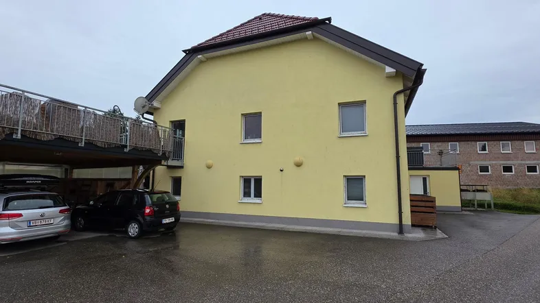 Gelbe Hausfassade mit Carport und gepflastertem Vorplatz unter bewölktem Himmel.