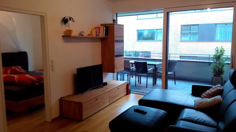 Helles Wohnzimmer mit Holzboden, gemütlichem Sofa und Zugang zu einem möblierten Balkon.