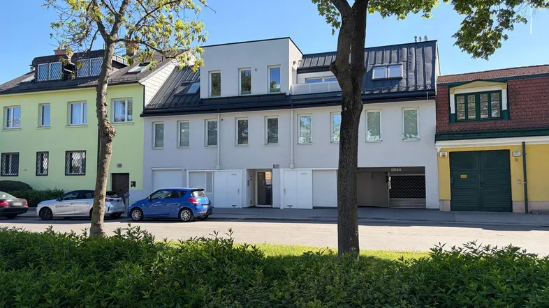 Modernes Mehrfamilienhaus mit heller Fassade, Garagen und parkenden Autos an einer begrünten Straße.