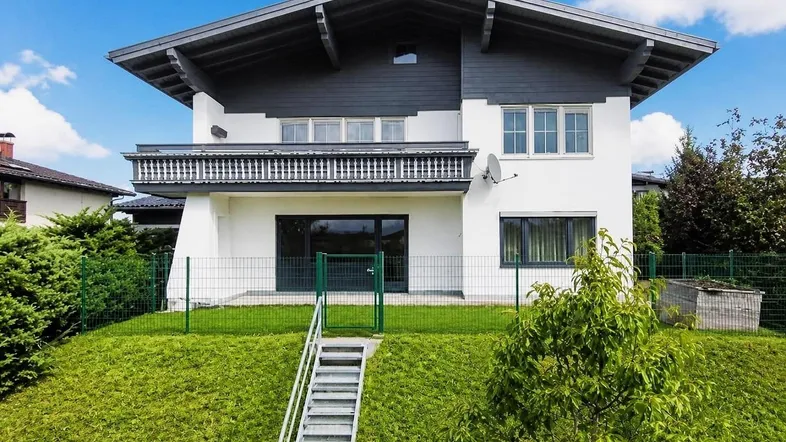 Modernes Einfamilienhaus mit weißer Fassade, dunklem Dach, Balkon und Gartenbereich.