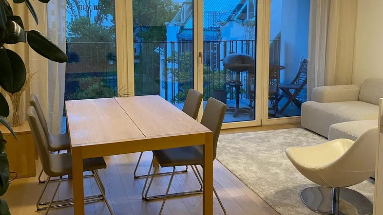 Heller Wohn- und Essbereich mit Holzboden, großem Fenster und Zugang zum Balkon.