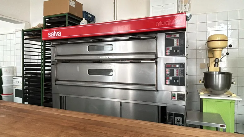 Große industrielle Öfen und ein Mixer in einer professionellen Bäckereiküche mit Holzarbeitsplatte und gefliesten Wänden.