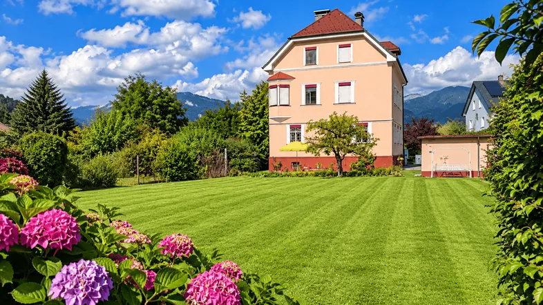 Haus mit großem, gepflegtem Rasen und blühenden Hortensien im Vordergrund, umgeben von Bäumen und Bergen.