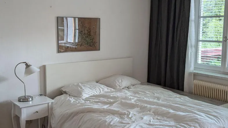 Helles Schlafzimmer mit Doppelbett, Nachttisch und Fenster mit dunklen Vorhängen.
