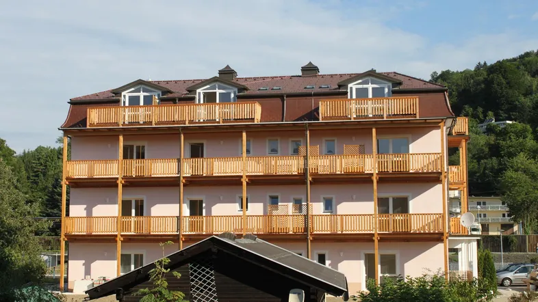 Mehrstöckiges Gebäude mit zahlreichen Holzbalkonen und Mansarddach, umgeben von viel Grün.