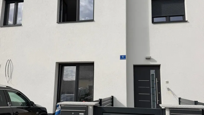 Moderne weiße Fassade eines Wohnhauses mit dunklen Fensterläden und Eingangstür, sowie einem geparkten Auto.
