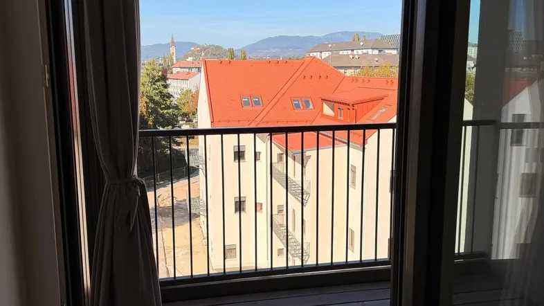 Blick von einem Innenraum auf einen Balkon mit Holzboden und Aussicht auf Dächer und Berge.