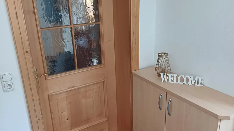 Heller Eingangsbereich mit Holztür, Fliesenboden und einem kleinen Schuhschrank.