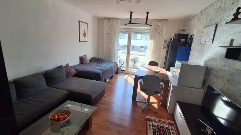 Gemütlicher Wohn- und Essbereich mit Schlafgelegenheit, Holzboden und Zugang zum Balkon.