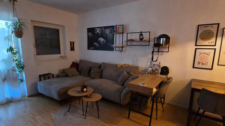 Gemütliches Wohnzimmer mit Ecksofa, Holzboden und einem Arbeitsbereich am Fenster.
