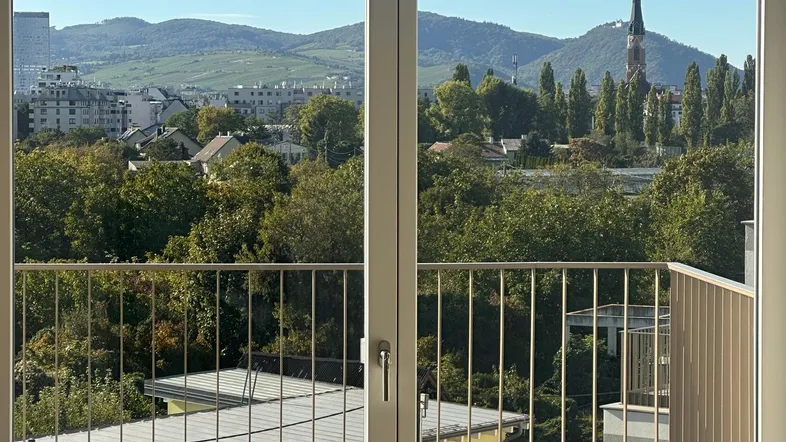 Heller Ausblick vom Balkon auf die Stadt, grüne Hügel und eine Kirche unter blauem Himmel.
