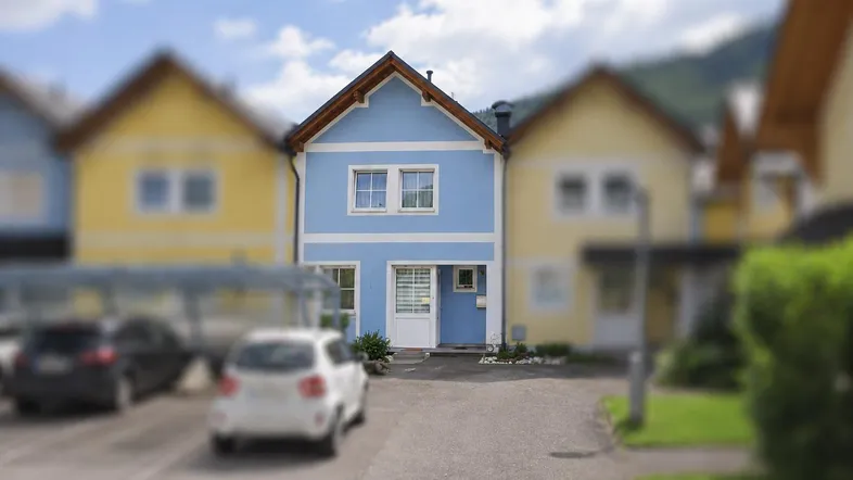 Blaues Reihenhaus mit weißer Fassadenverzierung und Parkplätzen davor in einer Wohnsiedlung.