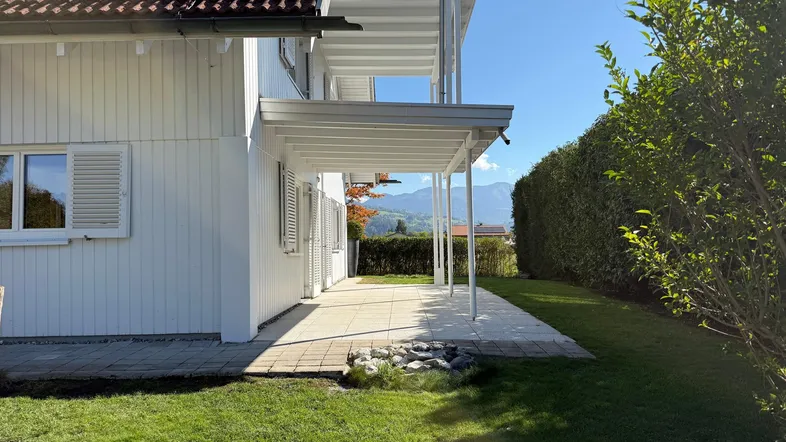 Helle Hausfassade mit überdachter Terrasse, gepflegtem Garten und Bergblick unter blauem Himmel.