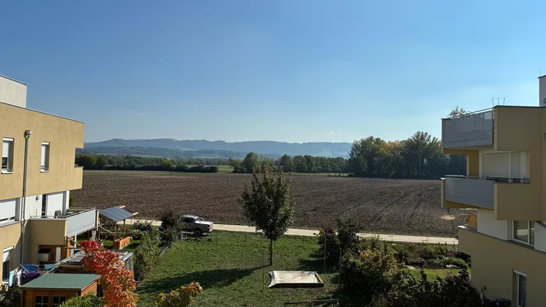 Weitblick über Felder und Berge von einem Balkon aus, mit angrenzenden Gebäuden und klarem Himmel.