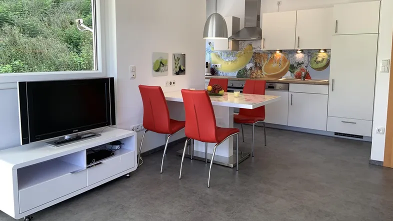 Heller offener Wohn- und Essbereich mit moderner Küche, Fernseher und Blick ins Grüne.