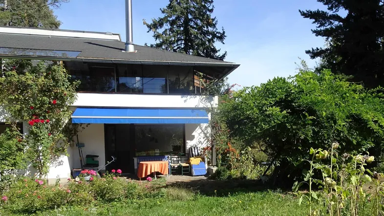 Hausfassade mit Terrasse, Garten und blauer Markise unter einem sonnigen Himmel.