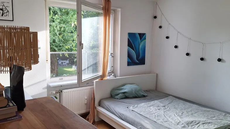 Helles Schlafzimmer mit Einzelbett, Schreibtisch und Blick ins Grüne durch das offene Fenster.