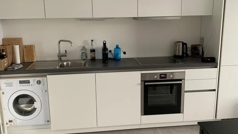 Moderne Einbauküche mit weißen Schränken, grauen Fliesen und integrierter Waschmaschine.