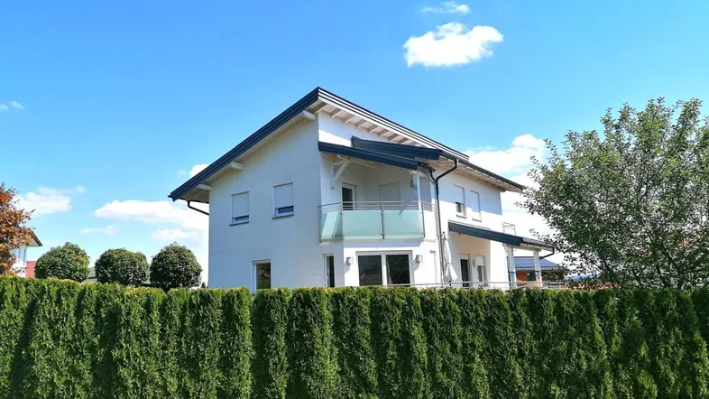 Modernes Einfamilienhaus mit Balkon und gepflegter Hecke unter blauem Himmel.